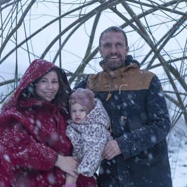 A family of three poses outdoors in a snowy winter scene. The parents smile warmly, while their bundled-up child looks curious amid softly falling snow.