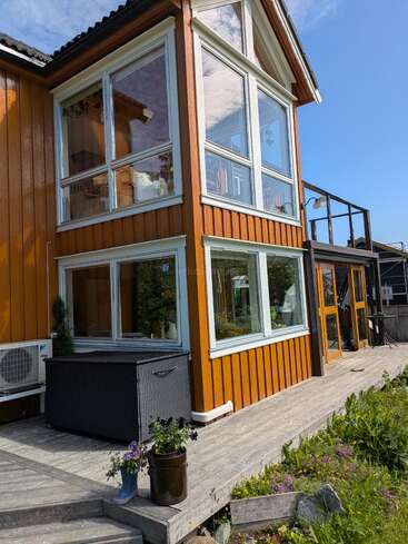 A modern wooden house with large glass windows, an outdoor wooden deck, flower pots, and clear blue skies, surrounded by greenery and vibrant blooming flowers.