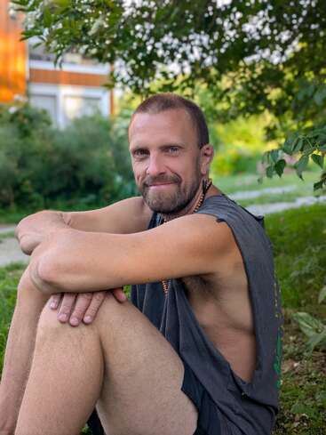 A man with short hair and beard sits outdoors on the grass. He is smiling, wearing a sleeveless shirt, with greenery and a house in the background.