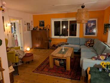 A cozy living room with warm lighting, orange accent wall, large L-shaped couch, wooden coffee table, eclectic decor, colorful rug, and a comfy reading chair.