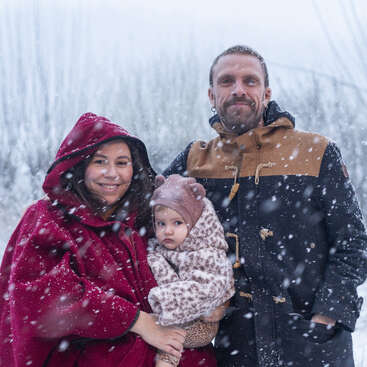 Una familia de tres miembros permanece unida bajo la nieve. Llevan abrigos de invierno. La madre sonríe con su bebé en brazos. El padre está a su lado, contento.