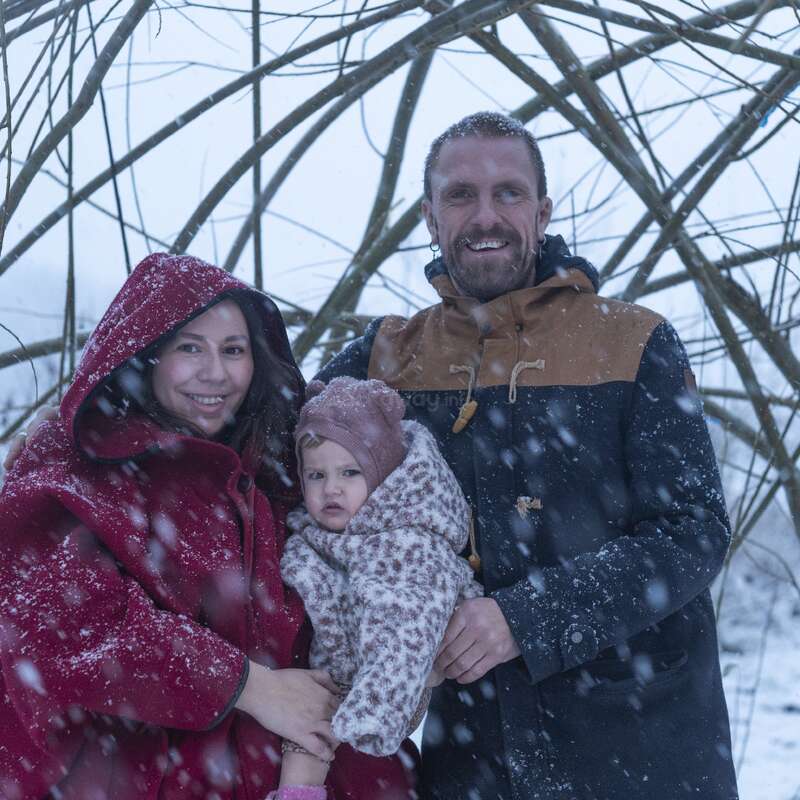 A family of three poses outdoors in a snowy winter scene. The parents smile warmly, while their bundled-up child looks curious amid softly falling snow.