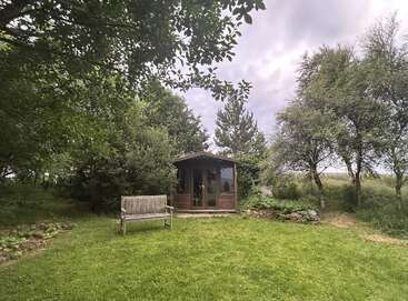 Das Bild zeigt eine ruhige Umgebung im Freien mit einem kleinen Holzschuppen, einer Bank und üppigem Grün vor einem bedeckten Himmel.