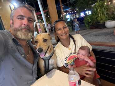 Un hombre, una mujer, un bebé y un perro sonrientes se sientan juntos en la mesa de un restaurante al aire libre por la noche. Ambiente animado con luces, plantas y gente de fondo.