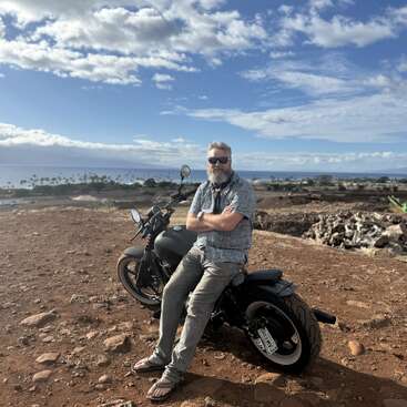 Un hombre con gafas de sol y barba está sentado en una motocicleta negra, en la cima de una colina rocosa, con vistas a las palmeras, el océano y un cielo parcialmente nublado.