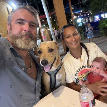 Un hombre, una mujer, un bebé y un perro sonrientes se sientan juntos en la mesa de un restaurante al aire libre por la noche. Ambiente animado con luces, plantas y gente de fondo.