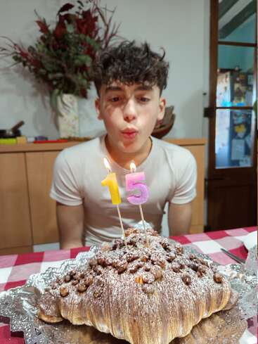 Un adolescente celebra su 15 cumpleaños soplando las velas de una tarta gigante de croissant en una mesa decorada, con un arreglo floral detrás.