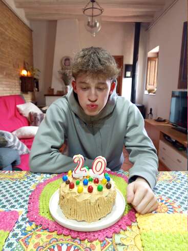 Un joven se sienta en una colorida mesa, soplando las velas de una tarta de cumpleaños decorada con caramelos y el número 20, en una acogedora habitación.