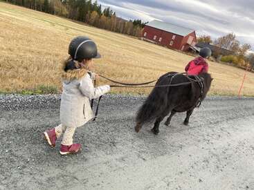 Two children, both wearing helmets, are outdoors. One rides a small black pony while the other leads it along a rural gravel road near fields and barns.