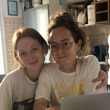 The image shows two women posing for a photo in a kitchen, with one woman\'s arm around the other, both wearing white t-shirts and smiling at the camera.