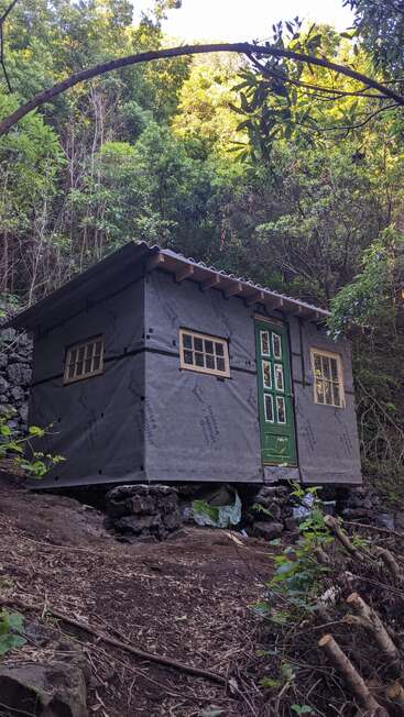 Une petite cabane inachevée est posée sur des blocs de pierre dans une forêt verdoyante. Elle possède une porte verte, des fenêtres simples et un toit en pente.