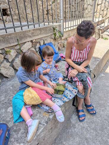 Eine Frau und zwei kleine Kinder sitzen auf Betonstufen im Freien und teilen sich eine Mahlzeit. Die Kinder tragen bunte Kleidung, und alle sehen entspannt und zufrieden aus.