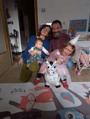 Eine glückliche Familie posiert in einem Haus. Die Mutter hält ein Baby, der Vater lächelt und ein aufgeregtes Mädchen mit einem Partyhut reitet auf einem bunten aufblasbaren Einhorn.