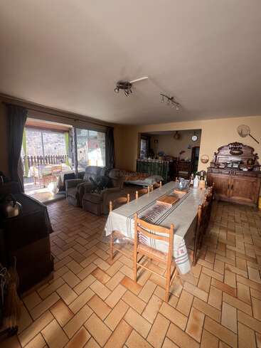This image shows a cozy dining and living area with a long table, wooden chairs, comfortable armchairs, large windows, tiled floor, and rustic wooden furniture.