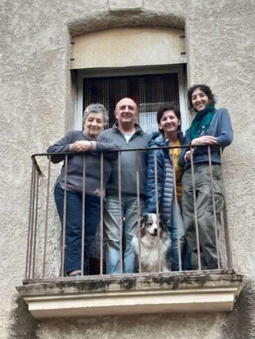 Cuatro personas y un perro posan en un pequeño balcón, sonriendo amablemente para la foto. El grupo parece relajado, disfrutando del tiempo que pasan juntos frente a un edificio rústico.
