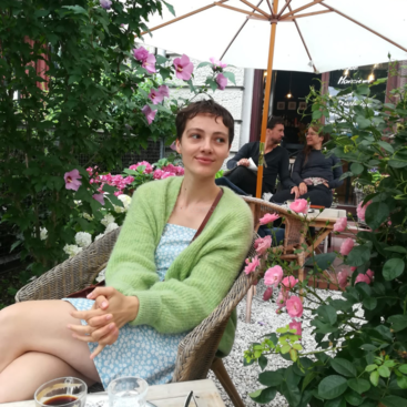 Das Bild zeigt eine Frau, die in einem Korbstuhl sitzt, umgeben von Blumen und Laub, mit einem Tisch vor ihr und Menschen im Hintergrund in einem Straßencafé.