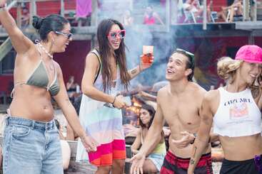 A group of young adults in swimwear and sunglasses are smiling, dancing, and enjoying themselves at an outdoor summer party or festival with a bonfire.