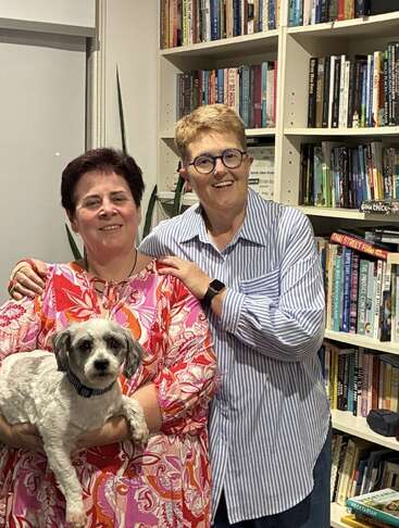 Zwei Frauen stehen zusammen in einem gemütlichen Raum voller Bücherregale und lächeln herzlich. Eine der Frauen hält einen kleinen Hund im Arm, was eine freundliche und einladende Atmosphäre schafft.
