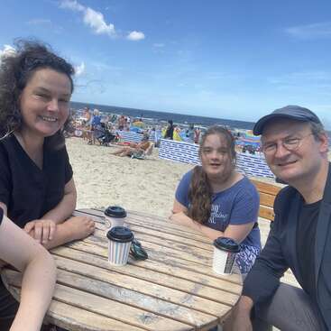 L'image montre une famille de trois personnes assise à une table en bois sur une plage, avec l'océan et le ciel bleu visibles en arrière-plan.