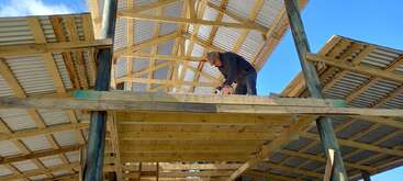 A worker is constructing a wooden building framework with a corrugated metal roof under a clear blue sky, using tools on the upper level of the structure.