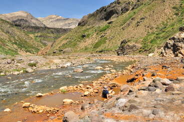 A person sits beside a rocky, mineral-rich stream in a mountainous valley. Green hills, exposed earth, and blue sky create a picturesque, remote natural landscape scene.