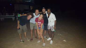Five friends pose playfully on a sandy beach at night. They are smiling and laughing, enjoying a fun, relaxed time together under the dark sky.