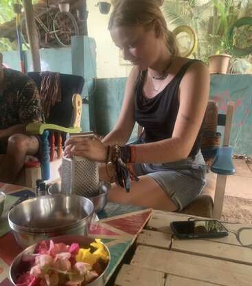 A young woman sits at a colorful table, grating food into a bowl. Flowers, metal bowls, and a phone rest nearby in a relaxed, tropical setting.