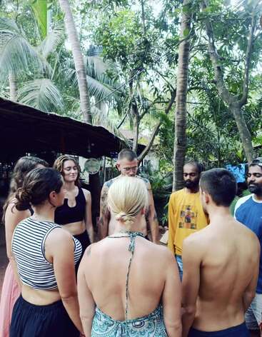 A group of people stands in a circle outdoors, surrounded by lush greenery and palm trees, appearing to meditate or engage in a peaceful group activity.