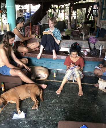 A group of people sit together indoors, writing in notebooks. Two dogs rest peacefully on the floor. The atmosphere is relaxed, cozy, and creatively focused.