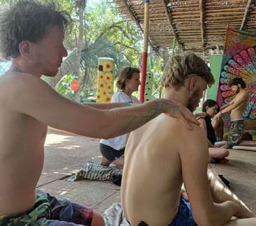 A group of people sit on the floor in a relaxed outdoor setting, giving each other shoulder massages. The atmosphere appears calm, peaceful, and communal.