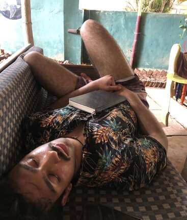 A man in a floral shirt is sleeping on a checkered couch, holding a closed book. He looks relaxed and comfortable, sprawled with eyes closed.