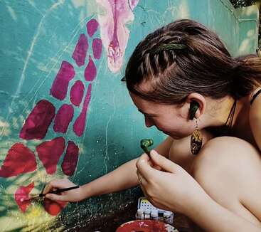 A young woman with braided hair is painting a colorful giraffe mural on a wall. She is focused, using pink paint, and wearing green earbuds.
