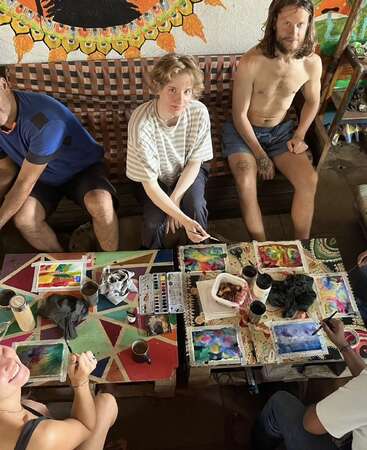 A group of people are gathered around colorful tables, painting vibrant watercolor artworks. They look relaxed and creative, surrounded by cups, snacks, and art supplies.