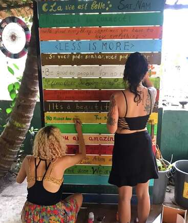 Two women are painting colorful inspirational quotes in various languages on wooden planks. They appear focused and creative, surrounded by plants and a peaceful outdoor setting.