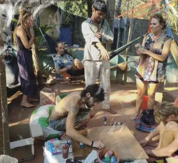 A group of people gather outdoors, engaging in creative activities like painting, drawing, and socializing. The scene feels relaxed, artistic, and community-focused, with vibrant surroundings.