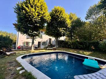 Un tranquilo jardín cuenta con una acogedora piscina, rodeada de frondosos árboles y vegetación. Al fondo, bajo un cielo azul y despejado, se alza una encantadora casa blanca.