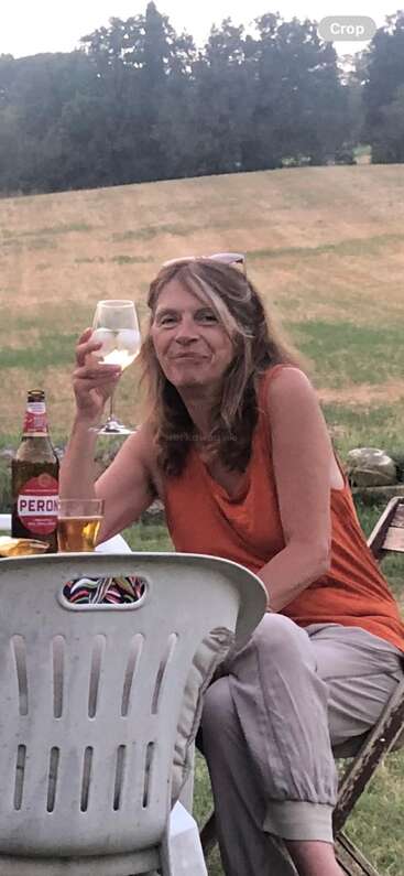 Una mujer de pelo largo está sentada al aire libre en una silla de plástico, sonriendo y con una copa de vino en la mano, rodeada de campos, árboles y una lata de cerveza Peroni.