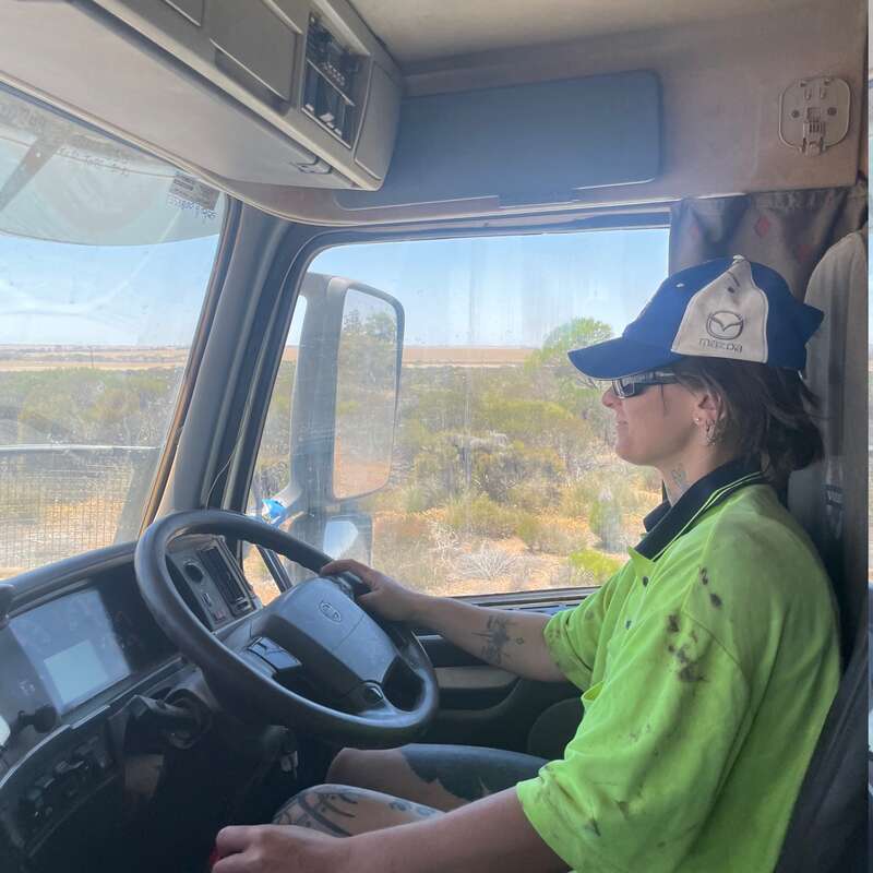 A person wearing a high-visibility shirt and Mazda cap is driving a truck through a rural landscape, focusing on the road ahead under bright daylight.