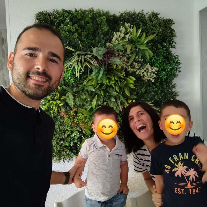 Une famille de quatre personnes souriante pose devant un mur végétal luxuriant. Les visages des deux enfants sont recouverts d'autocollants emoji souriants afin de préserver leur intimité.