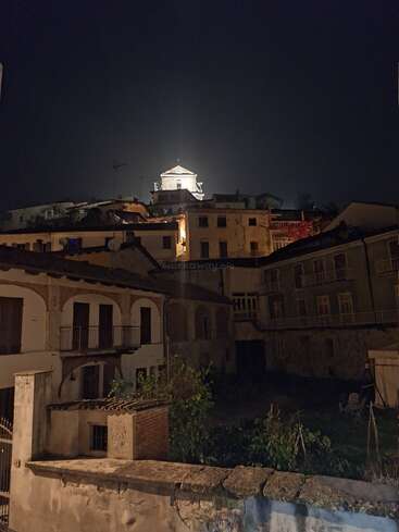 Uma antiga vila europeia à noite, iluminada por luzes aconchegantes, com um edifício imponente brilhando no topo da colina sob um céu escuro e claro. Atmosfera tranquila e serena.