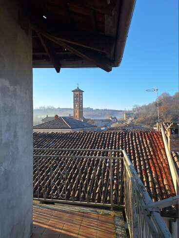 Uma varanda encantadora tem vista para os telhados de terracota e uma torre do relógio ao longe. O orvalho brilha sob um céu azul límpido, enquanto a luz da manhã ilumina o cenário tranquilo e pitoresco da vila.