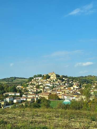 Uma pitoresca vila no topo de uma colina, cercada por uma vegetação exuberante sob um vasto céu azul, apresenta casas charmosas agrupadas em torno de uma igreja central, evocando uma atmosfera tranquila e idílica.