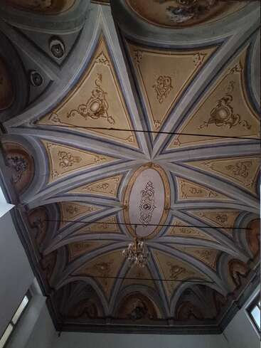 Esta imagem mostra um teto abobadado e ornamentado, com desenhos dourados intricados, padrões geométricos e um lustre no centro. A arquitetura parece clássica, transmitindo uma sensação de elegância.
