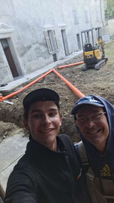 Dois homens sorridentes tiram uma selfie em um canteiro de obras, com tubos laranja, uma pequena escavadeira, terra e um grande prédio cinza ao fundo.