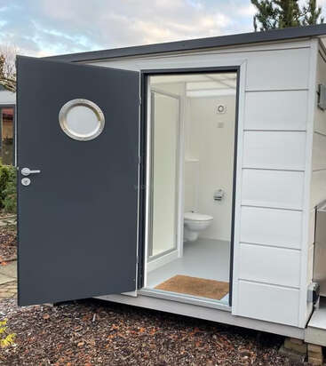 Um pequeno e moderno banheiro externo branco com uma porta cinza escuro, uma janela circular e um vaso sanitário visível no interior, situado em uma área de jardim com cobertura vegetal e vegetação.