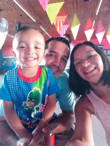 The image shows a family of three posing for a selfie in a room with a wooden ceiling and colorful triangular flags hanging from it.