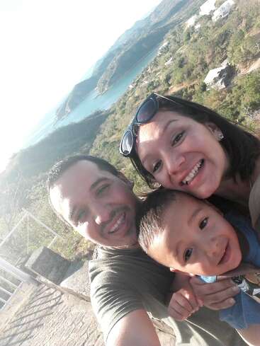 The image depicts a family of three posing for a selfie in front of a scenic landscape, with a body of water and mountains visible in the background.