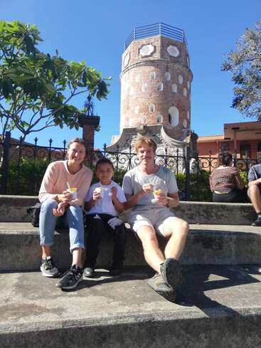 The image depicts a family of three sitting on concrete steps, enjoying ice cream cones in front of a historic brick tower on a sunny day.