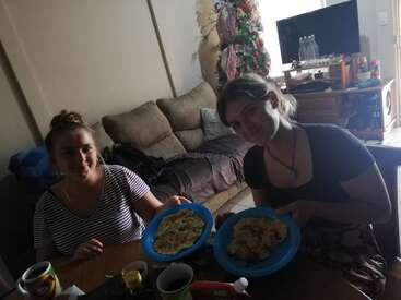 The image depicts two women sitting at a table, each holding a plate of food, with a Christmas tree and TV visible in the background of their living room.