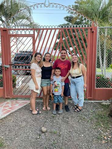 The image depicts a family of five standing in front of a red gate, posing for a photo, with a serene background of trees and a blue sky.